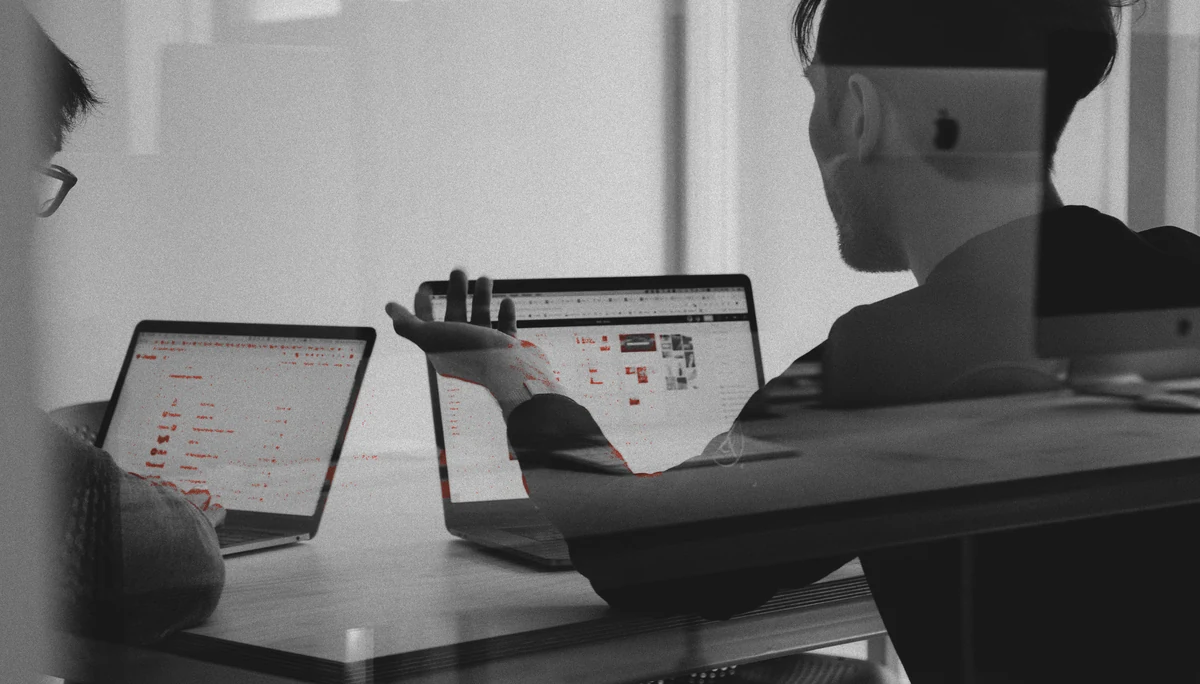 Two developers at a shared desk, one explaining a change over open laptops